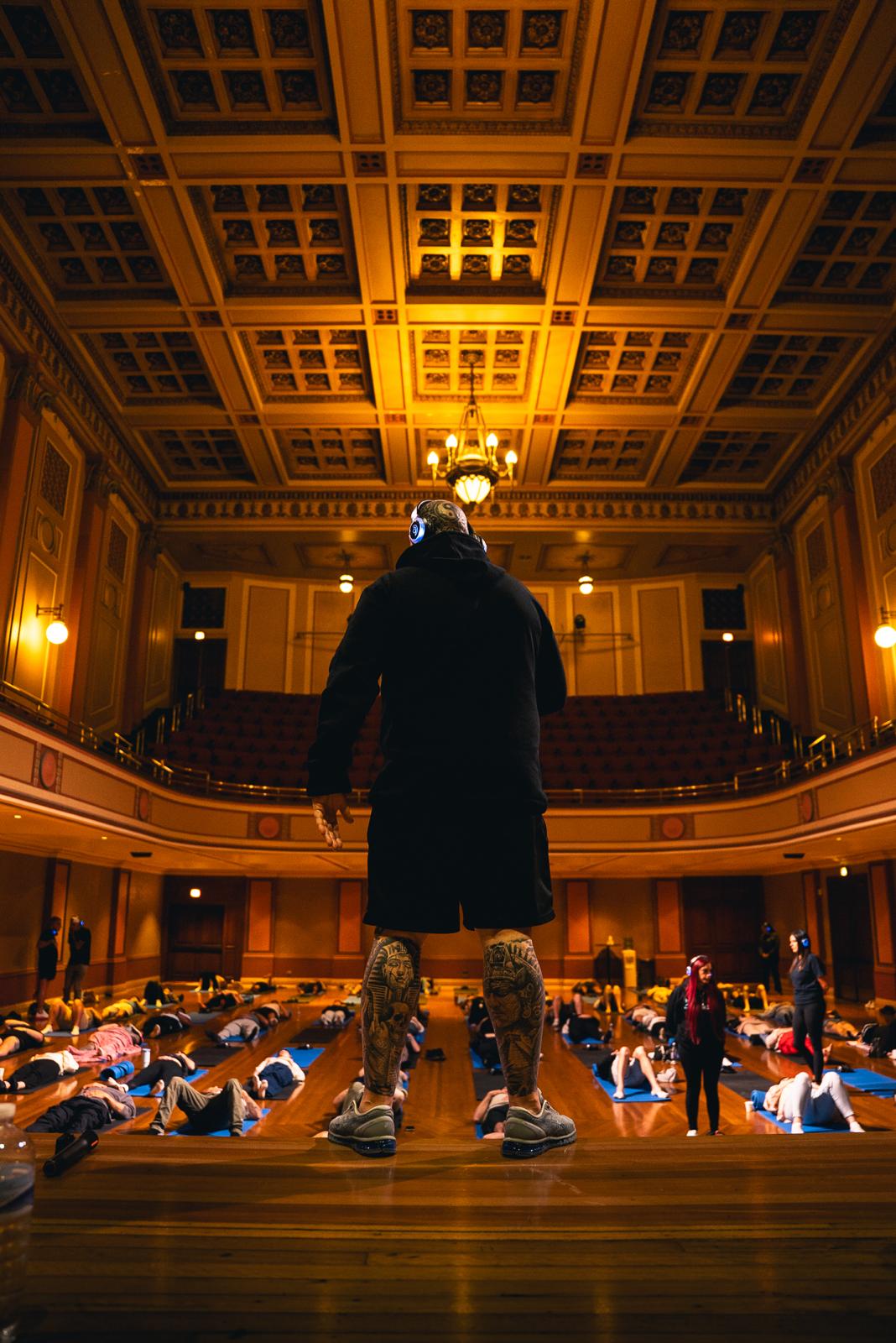 Gaz Wright standing on stage in a grand hall, facing rows of attendees lying on mats