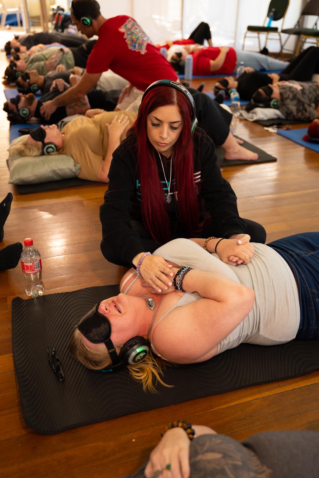 Facilitator kneeling beside a participant resting on a mat with headphones and eye mask
